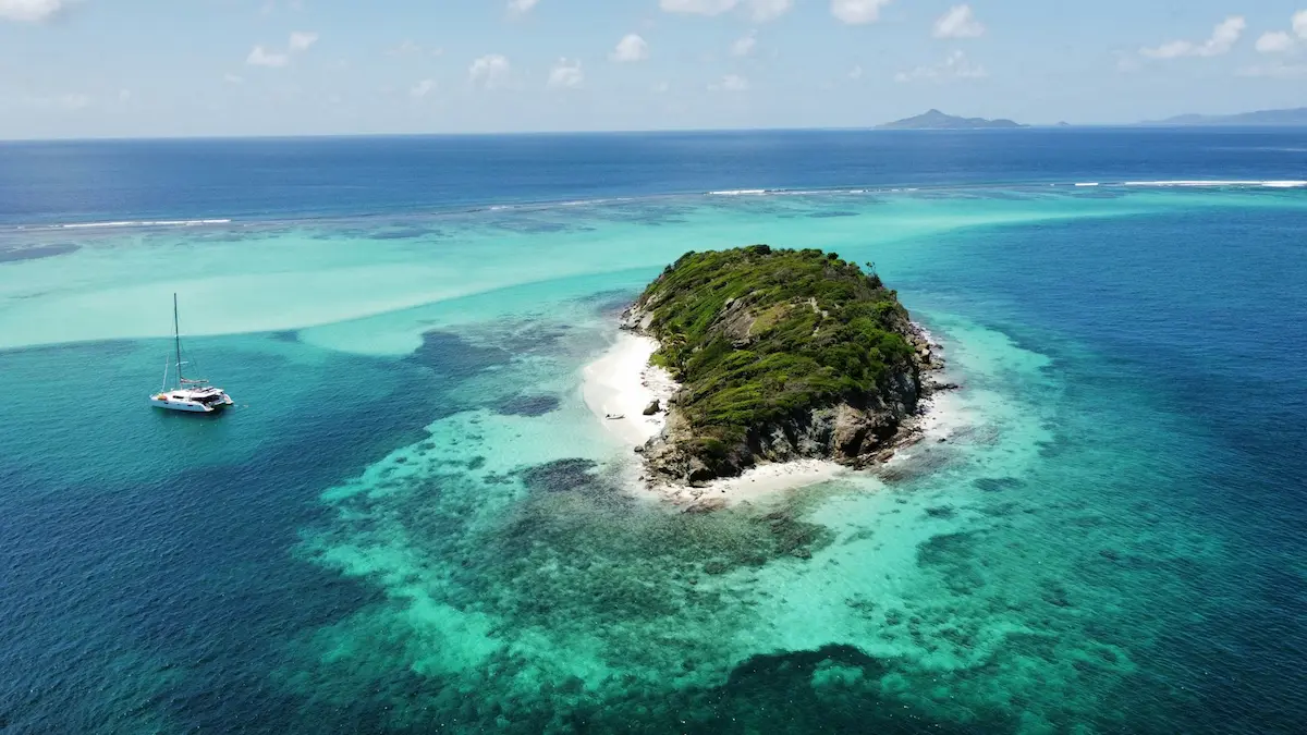 Small tropical island in the Tobago Cays surrounded by turquoise water with a catamaran anchored nearby under clear Caribbean skies.