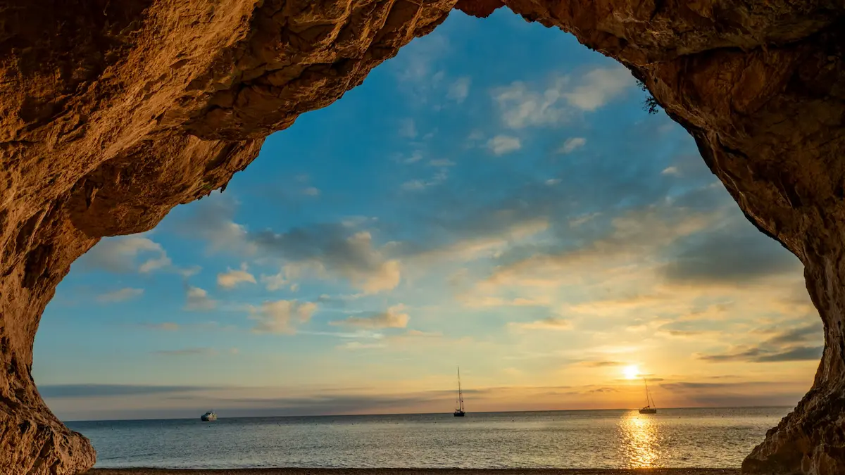 View through a sea cave to sailboats at sunset off Cala Luna, Sardinia