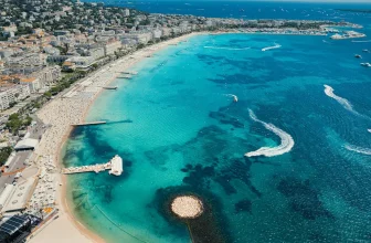 Aerial view of a long sandy beach and turquoise bay on the French Riviera with yachts, piers, and Mediterranean resort buildings.