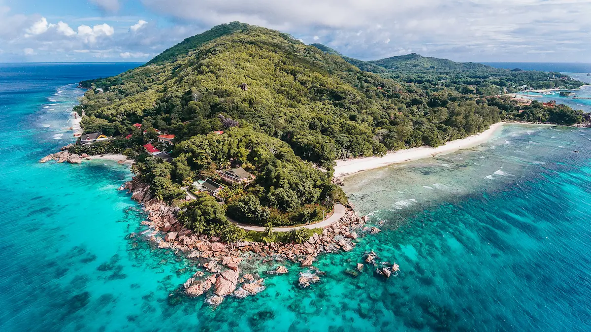 Aerial view of La Digue, Seychelles with granite rocks, white-sand beaches, turquoise water, lush vegetation, and coastal buildings near the shoreline.