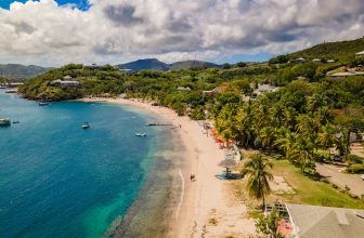 White-sand beach in Antigua with turquoise water, anchored sailboats, palm trees, and coastal homes under a partly cloudy Caribbean sky.